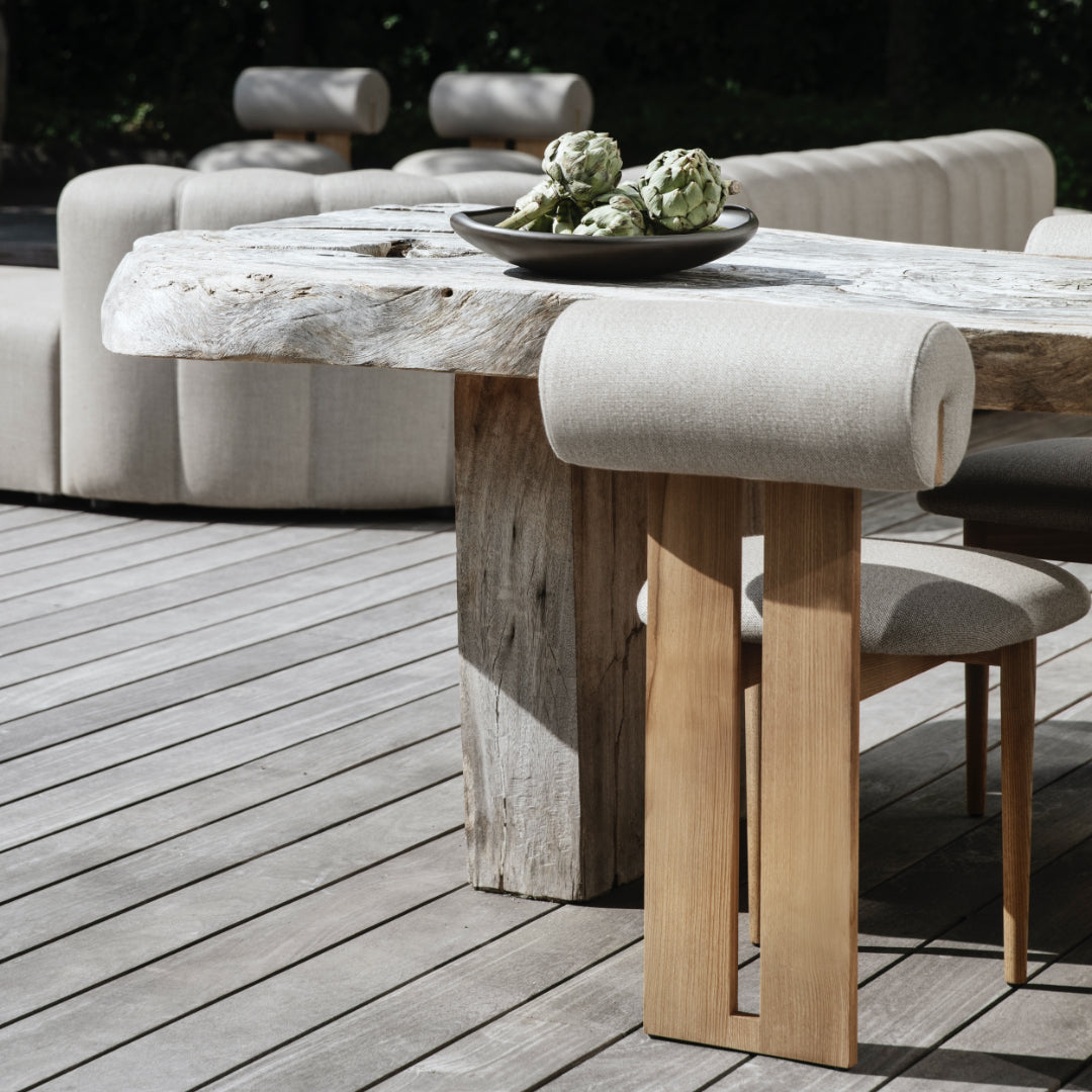 Wooden dining table with natural finish on a wooden deck, surrounded by cushioned chairs.