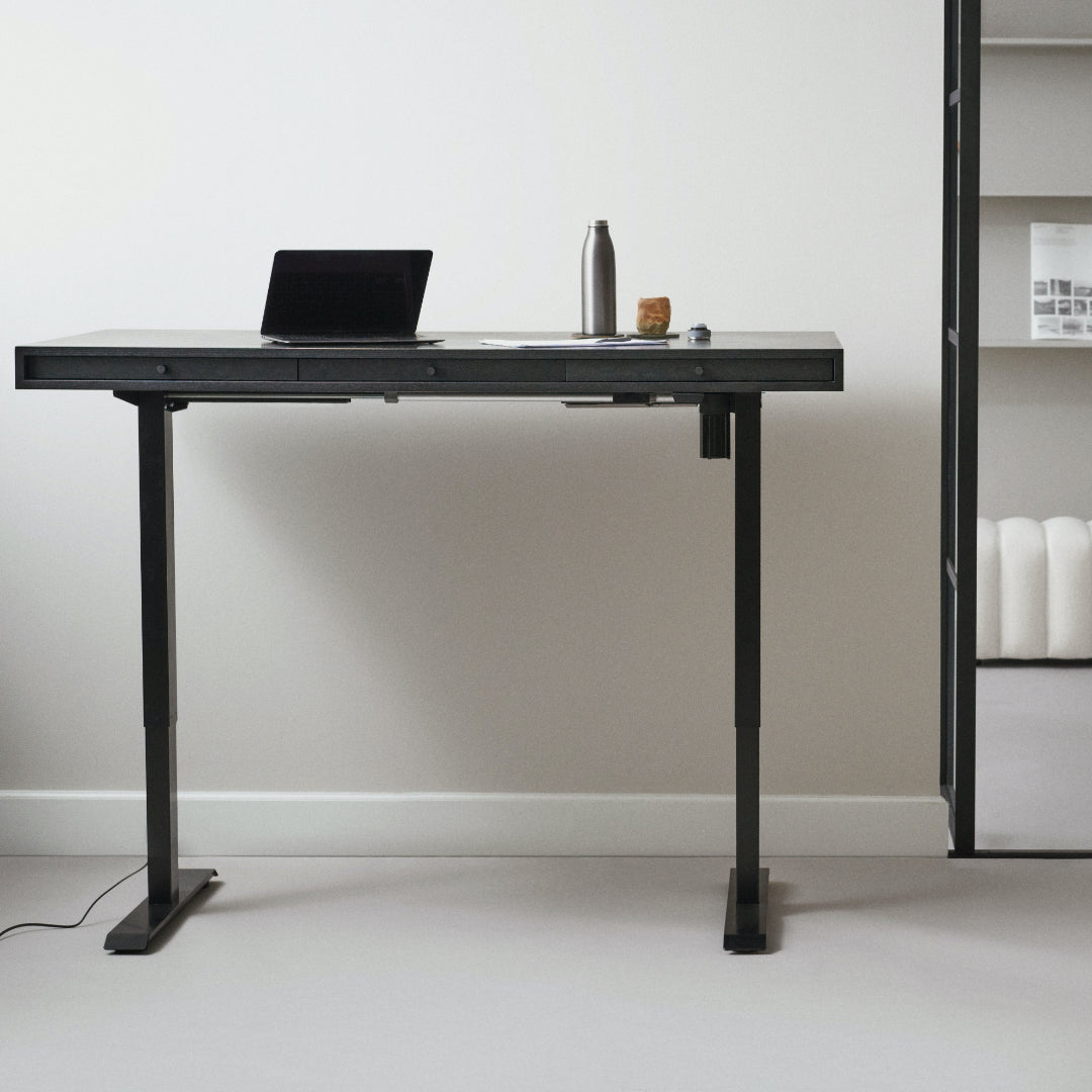 Black adjustable sitting/standing desk with a laptop, water bottle, and mug on a white wall background - NORR11 Home Desk