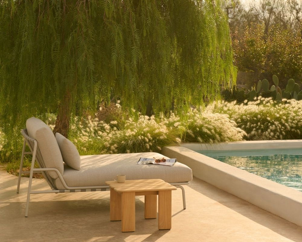 A light beige outdoor chaise lounge with matching cushions sits beside a swimming pool on a sunlit patio, with a small wooden side table in front. A large leafy tree and landscaped garden are visible in the background.